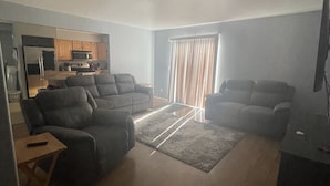 A living room with two sofas, a coffee table, and a TV stand. There is a kitchen area with cabinets and appliances in the background.
