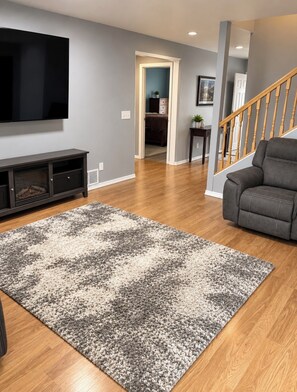 Un salon doté d’un téléviseur à écran plat, d’un meuble de rangement en bois foncé, d’un tapis gris et blanc, d’un canapé gris et d’un escalier en bois.