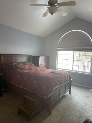 A bedroom with a large bed, a ceiling fan, a dresser, and a window with a view of trees.
