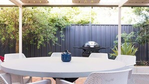 Una veranda coperta con un tavolo rotondo e sedie in rattan bianco, un barbecue e un vaso azzurro.