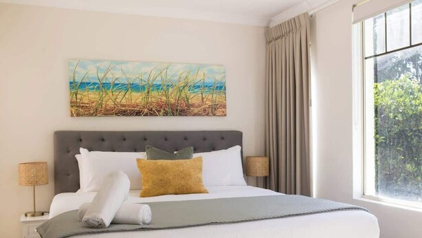 Un letto rifatto con cura, con una testiera grigia, un quadro raffigurante una spiaggia sopra e una finestra con tende a destra.