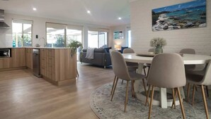 Una cucina moderna con mobili in legno e lavastoviglie integrata, una zona pranzo con tavolo e sedie bianche, e un quadro raffigurante una spiaggia.