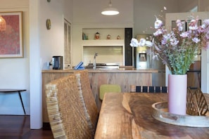 A wooden dining table with a vase of flowers, a bench, and a kitchen area with a white countertop and shelves. - Higgins Hideaway—Central Cottage with Garden Oasis (Margaret River)