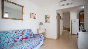 Una sala de estar con sofá, una mesita de noche con lámpara, y un área de cocina con refrigerador y armarios.