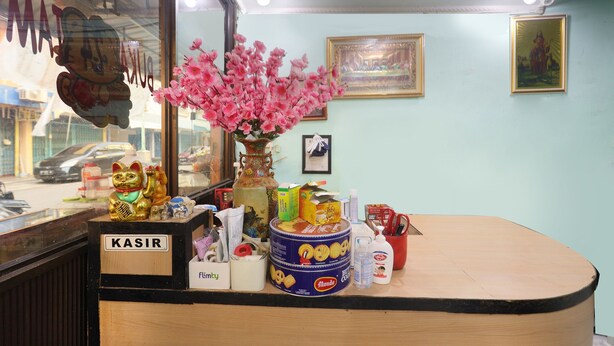 Un mostrador de recepción con un jarrón de flores rosadas, una estatua de un gato dorado y varios objetos, incluyendo una caja con la etiqueta 'KASIR'.