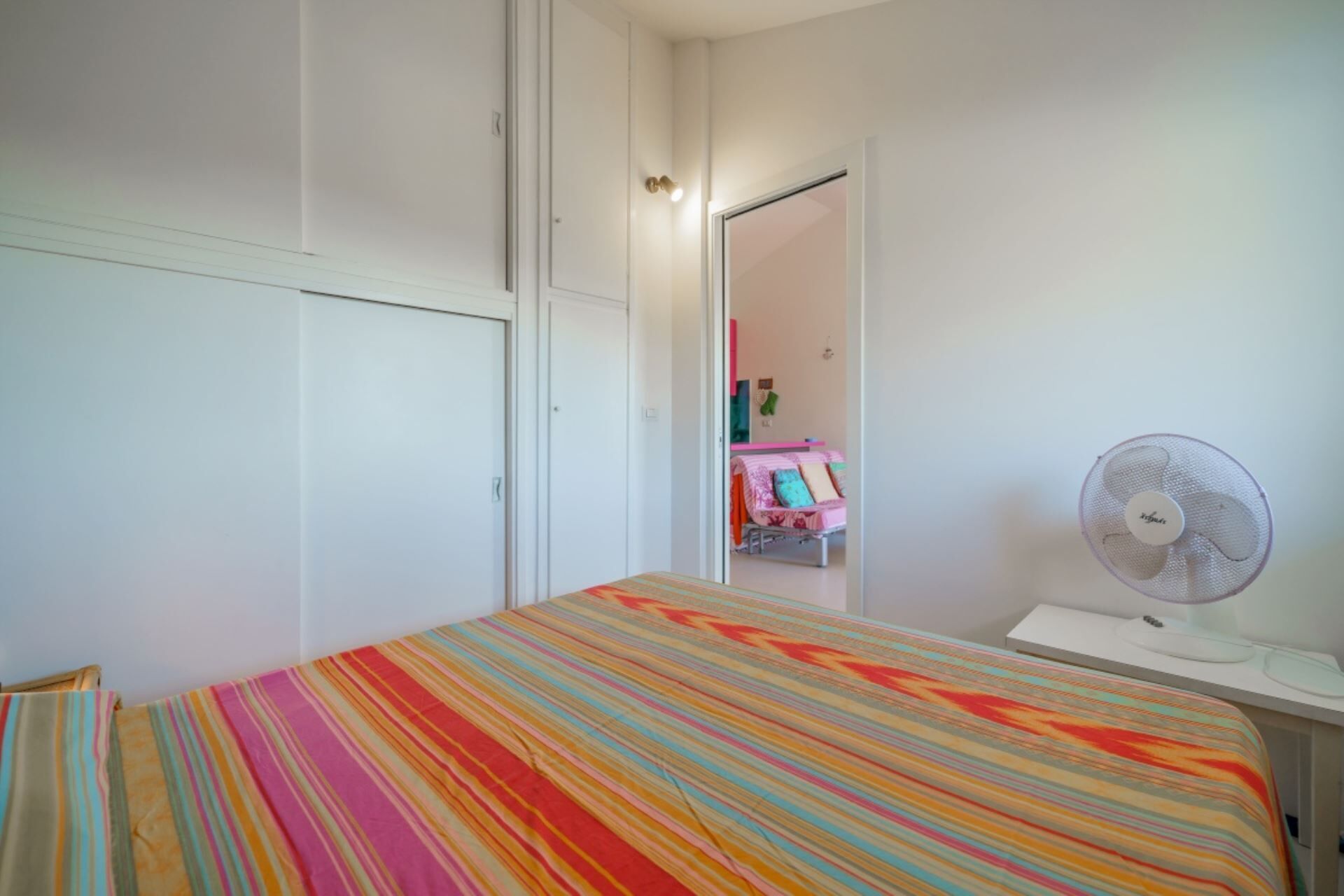 Een slaapkamer met een bed, een wit bureau met een ventilator en een deur naar een andere kamer.