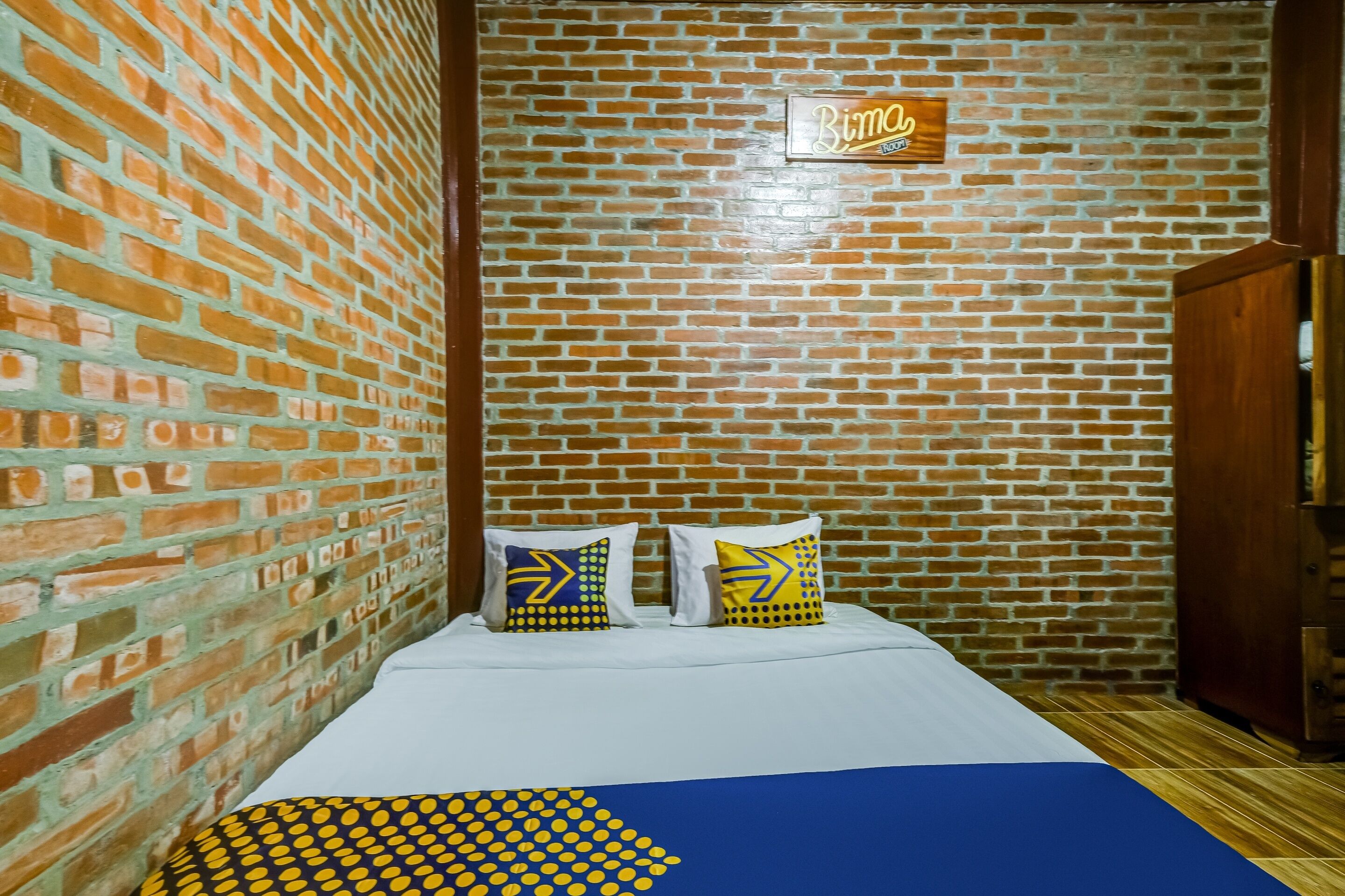 Una habitación con pared de ladrillo, una cama con ropa de cama blanca, dos almohadas con diseños geométricos y un cartel sobre la cama.