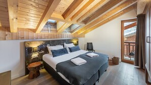 A bedroom with a wooden ceiling, a large bed, a headboard with artwork, a chair, and a view of the outside through a window. - MONT BLANC - A08 - apartment for 8 people with 3 bedrooms (LA CHAPELLE D ABONDANCE)