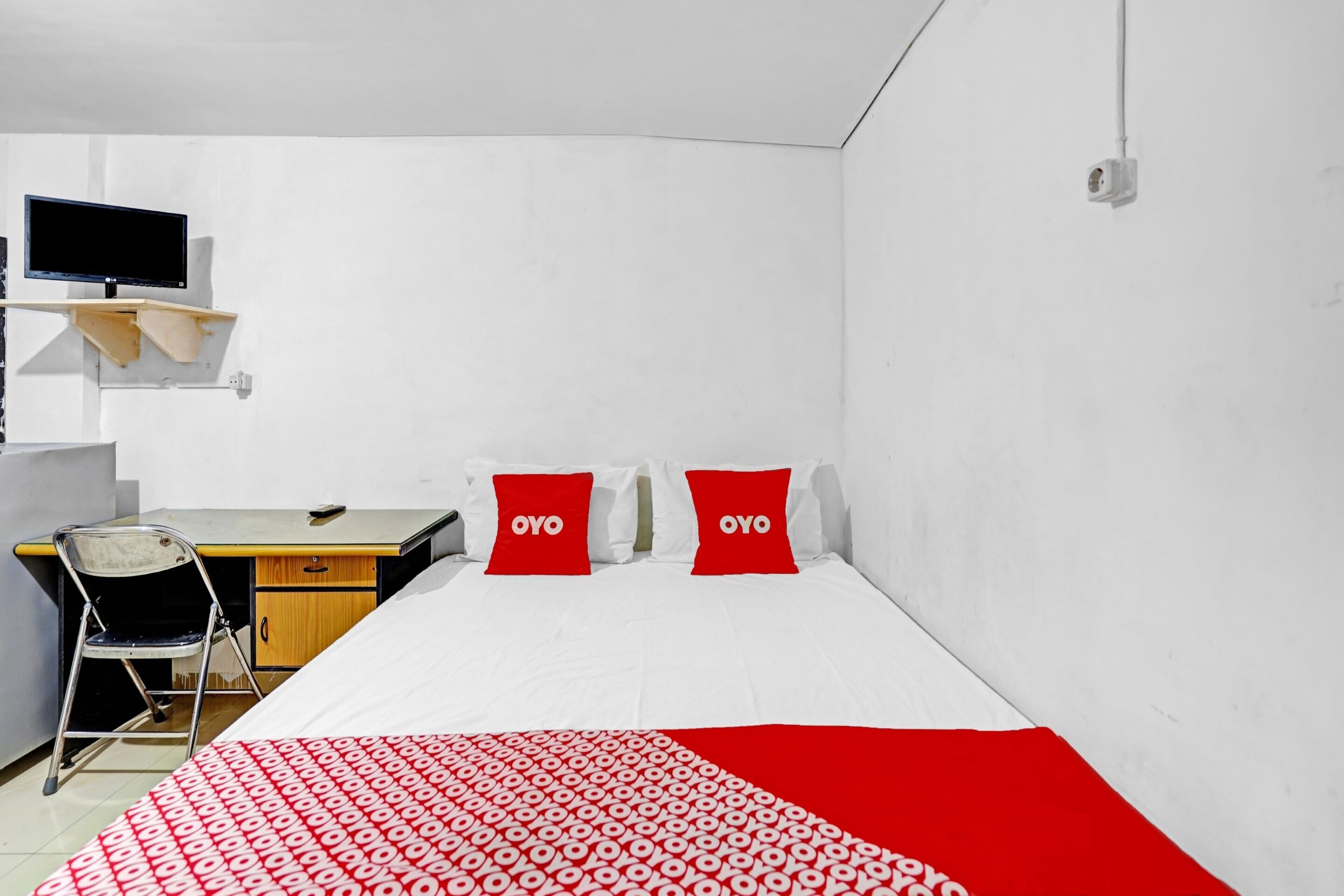 A room with a bed, two red pillows, a desk with a chair, and a television on the wall.