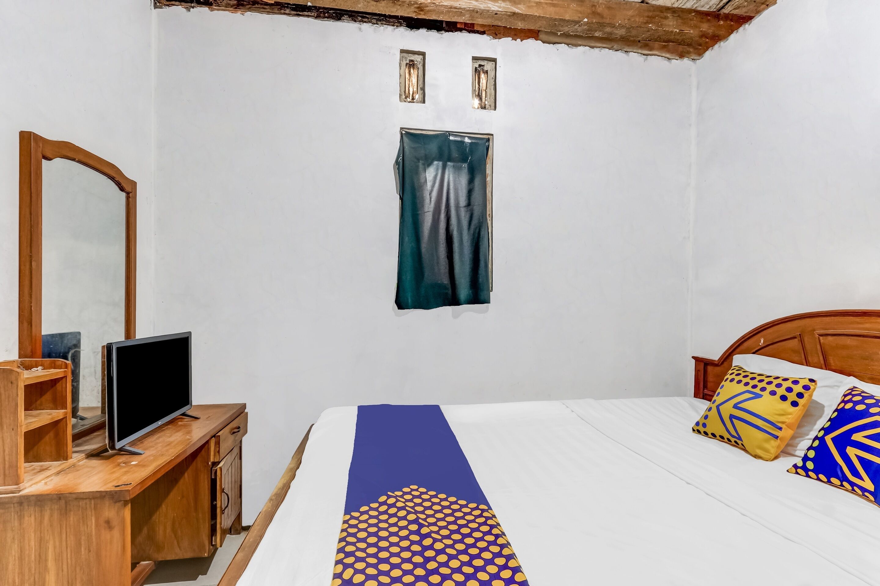 Une chambre avec un bureau en bois, une télévision, un lit avec des coussins à motifs, une fenêtre avec un rideau et un miroir sur le mur.
