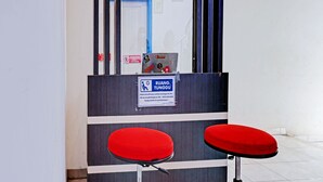 A reception area with a desk and two red cushioned stools. A sign above the desk reads "RUANG TUNGGU" with contact information. - Hotel O Bumi Merpati Residence (Makassar)