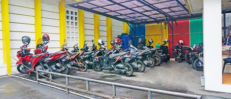Un estacionamiento para motocicletas con un techo cubierto, una barrera de cadena y fachada de edificio amarillo y blanco.