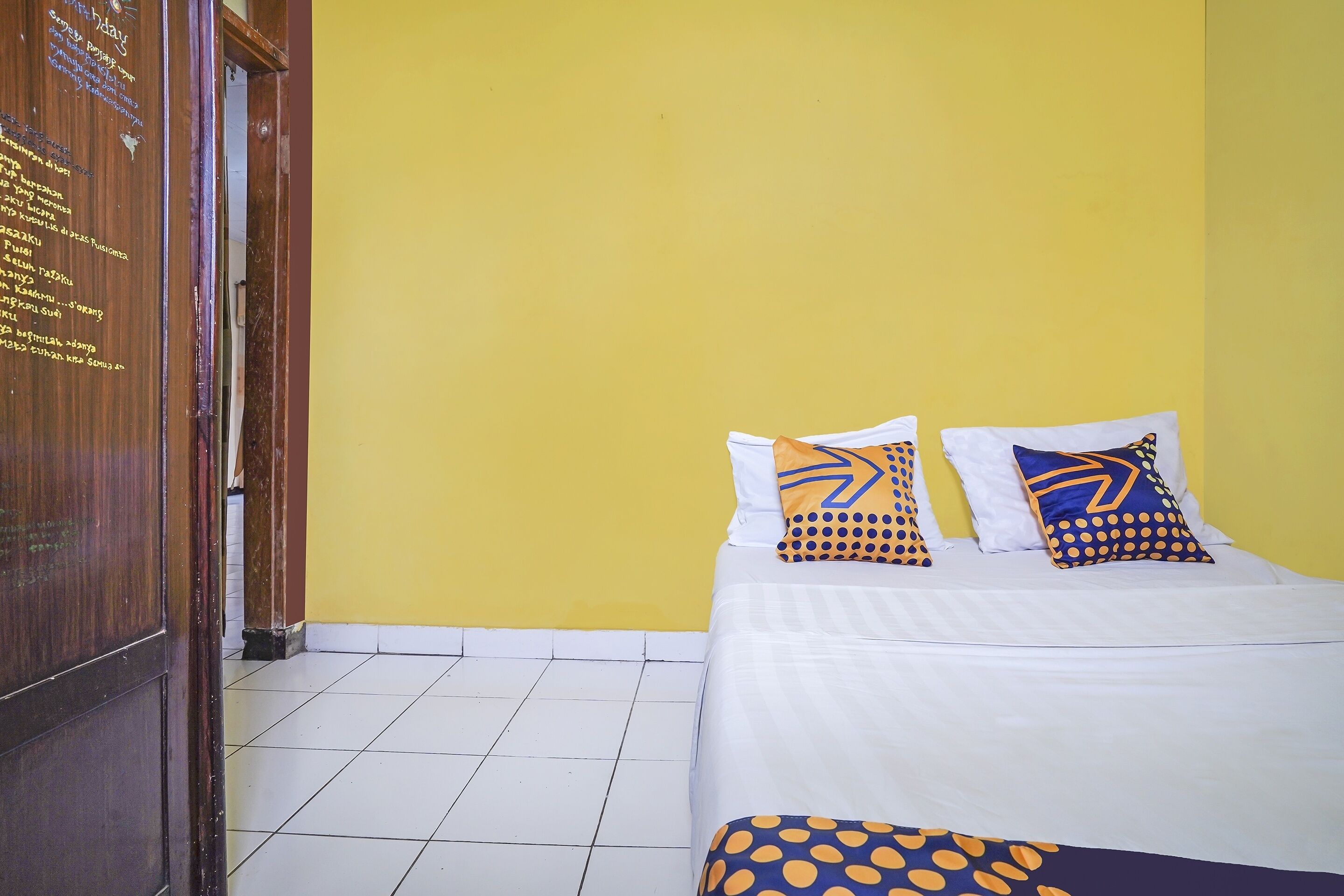 Une chambre d’hôtel avec un lit, deux oreillers à motifs géométriques, une porte en bois avec des inscriptions et un mur jaune.