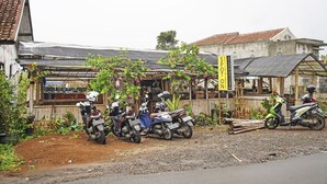 Pequeña tienda a un lado del camino, con un letrero, algunas scooters estacionadas y suelo de tierra.