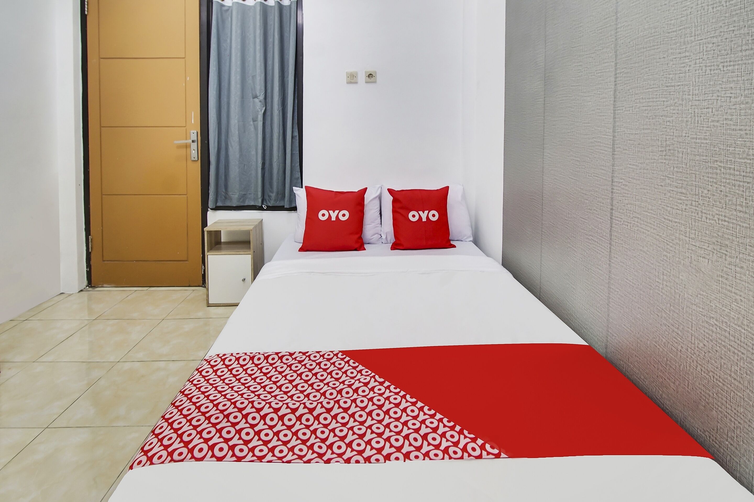Habitació d'hotel amb un llit, dos almoinos vermells, un edredó blanc i vermell amb dibuixos, una porta de fusta, una finestra amb cortines i una paret blanca.