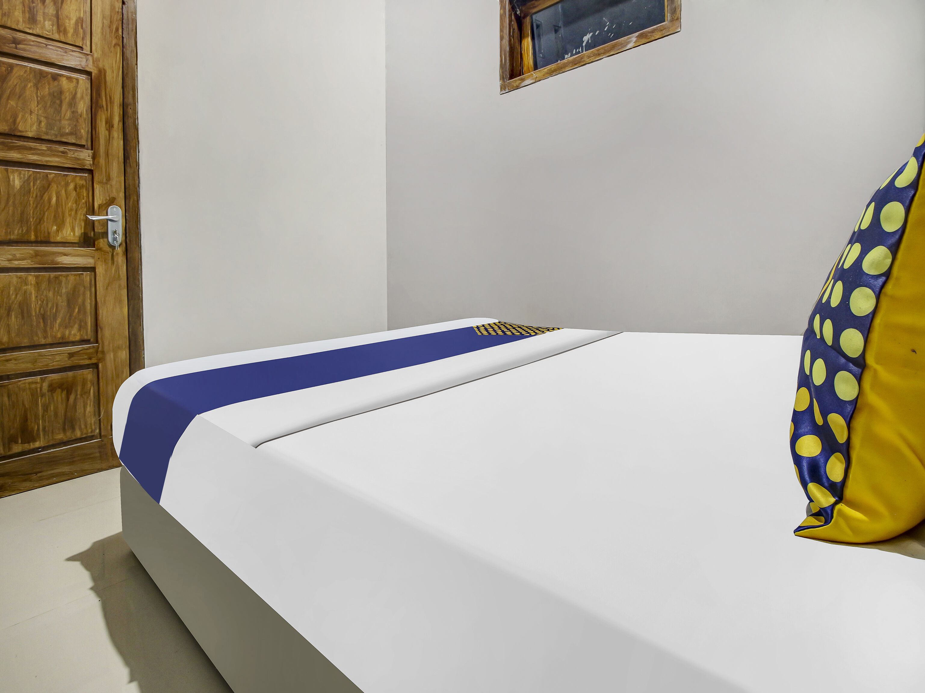 Uma cama bem arrumada com colcha azul e branca, uma porta de madeira e uma pequena janela com moldura de madeira.