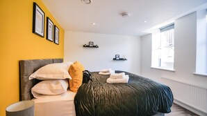 Un dormitorio con cama, almohadas, mesita de noche, lámpara y ventana.