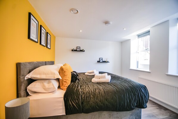 A bedroom with a bed, pillows, a bedside table, a lamp, and a window.