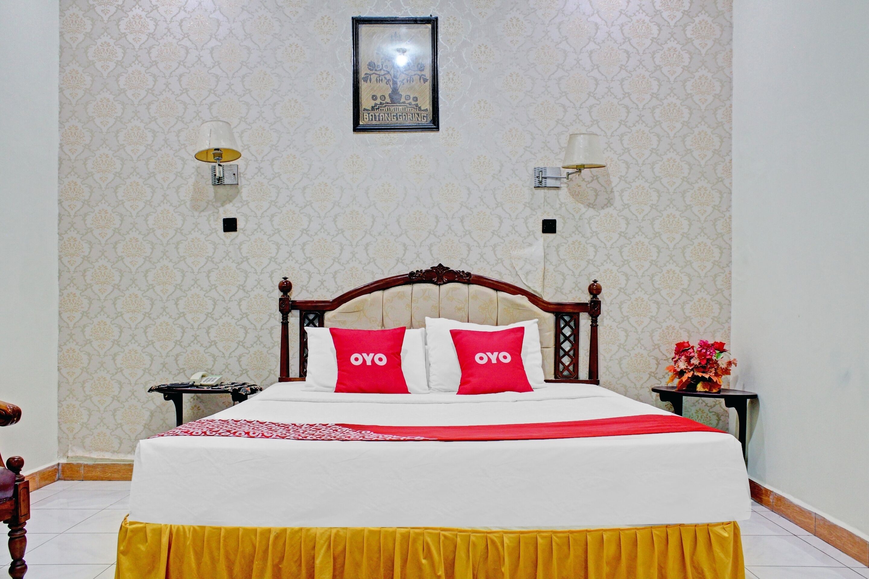 A neatly made bed with a wooden headboard, two pillows with "OYO" on them, and a red and white bedspread. There are two wall-mounted lamps and a framed picture above the bed.