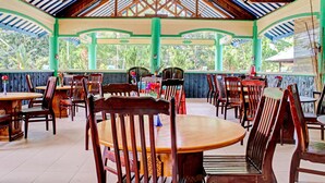 A covered outdoor dining area with wooden tables and chairs, surrounded by a green and blue color scheme.