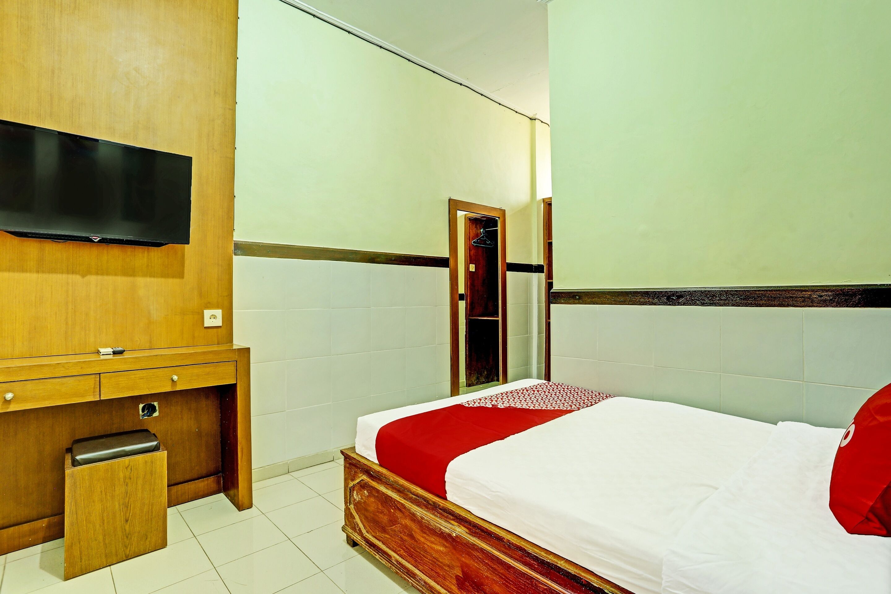A hotel room with a wooden desk, a television, a bed with a red and white bedspread, and a mirror on the wall.