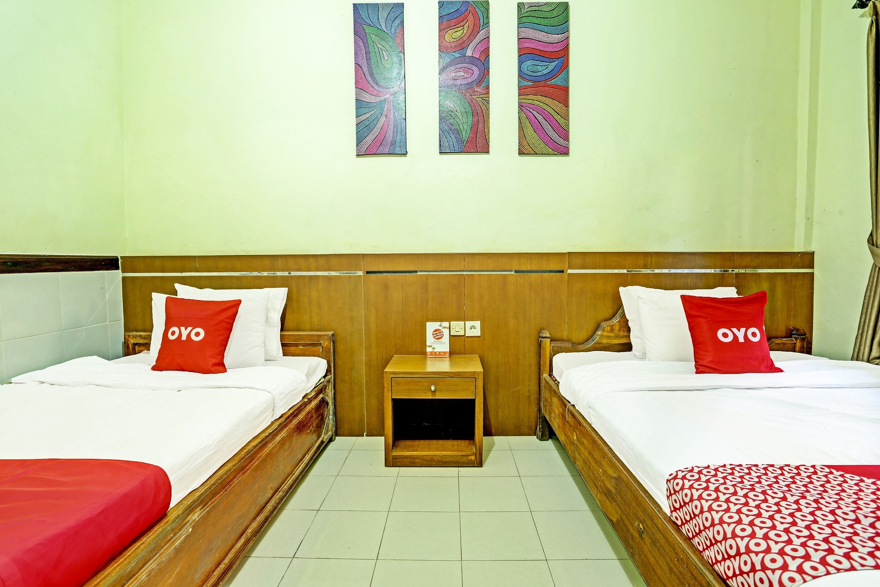Une chambre d’hôtel avec deux lits, chacun doté d’une tête de lit en bois, de draps blancs et d’oreillers rouges ornés du mot « OYO ». Deux tableaux abstraits sont accrochés au mur au-dessus des lits.