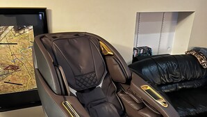 A massage chair with a control panel, positioned in a living room with a wooden floor and a black leather sofa. - Unique spacious upper Condo Suite. Steps from waterfront  (Sarnia)