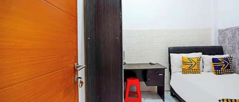 A bedroom with a wooden door, a bed with patterned pillows, a small desk, and a red stool.
