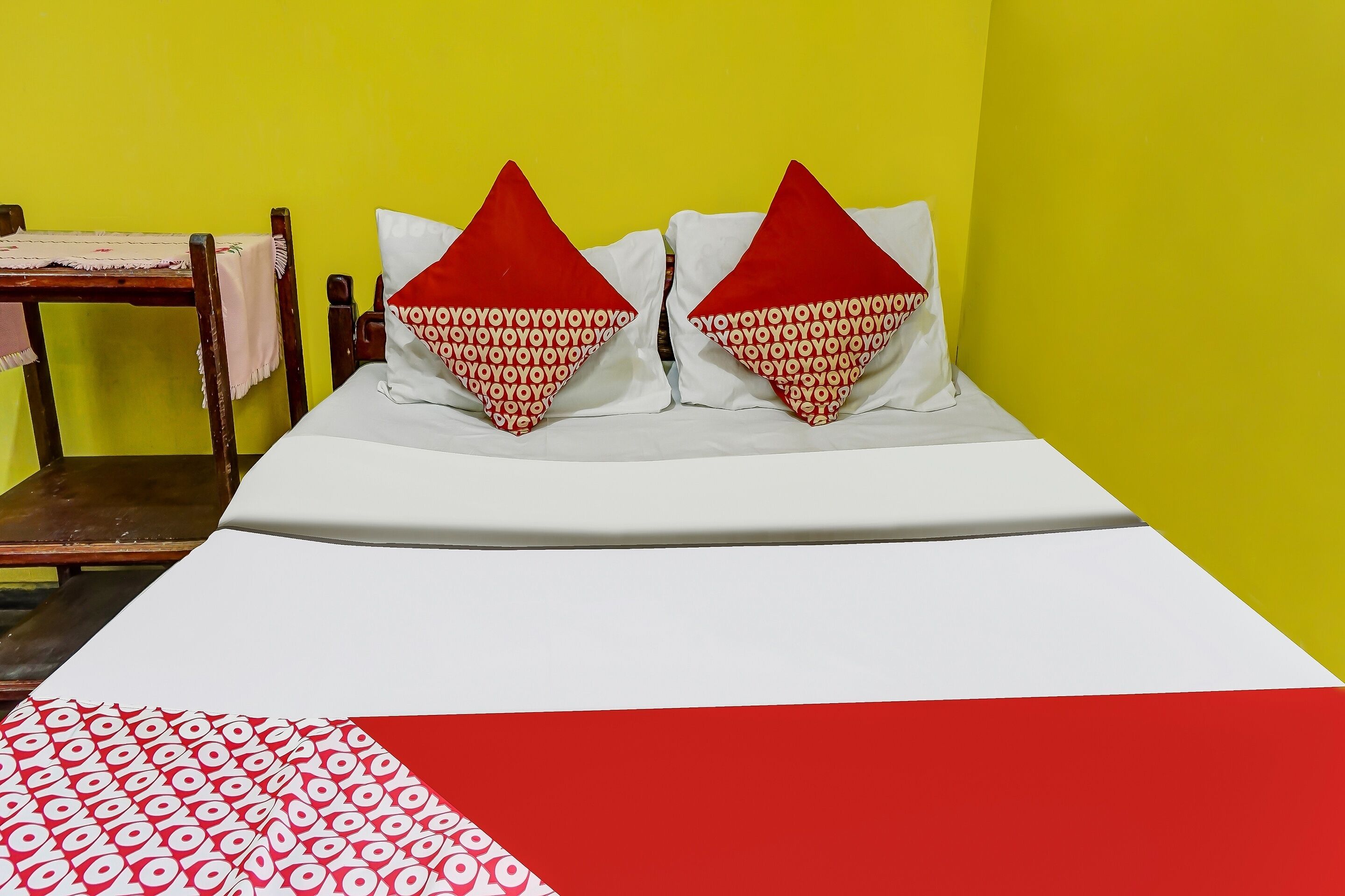 A bed with two pillows, a wooden chair, and a table against a yellow wall.