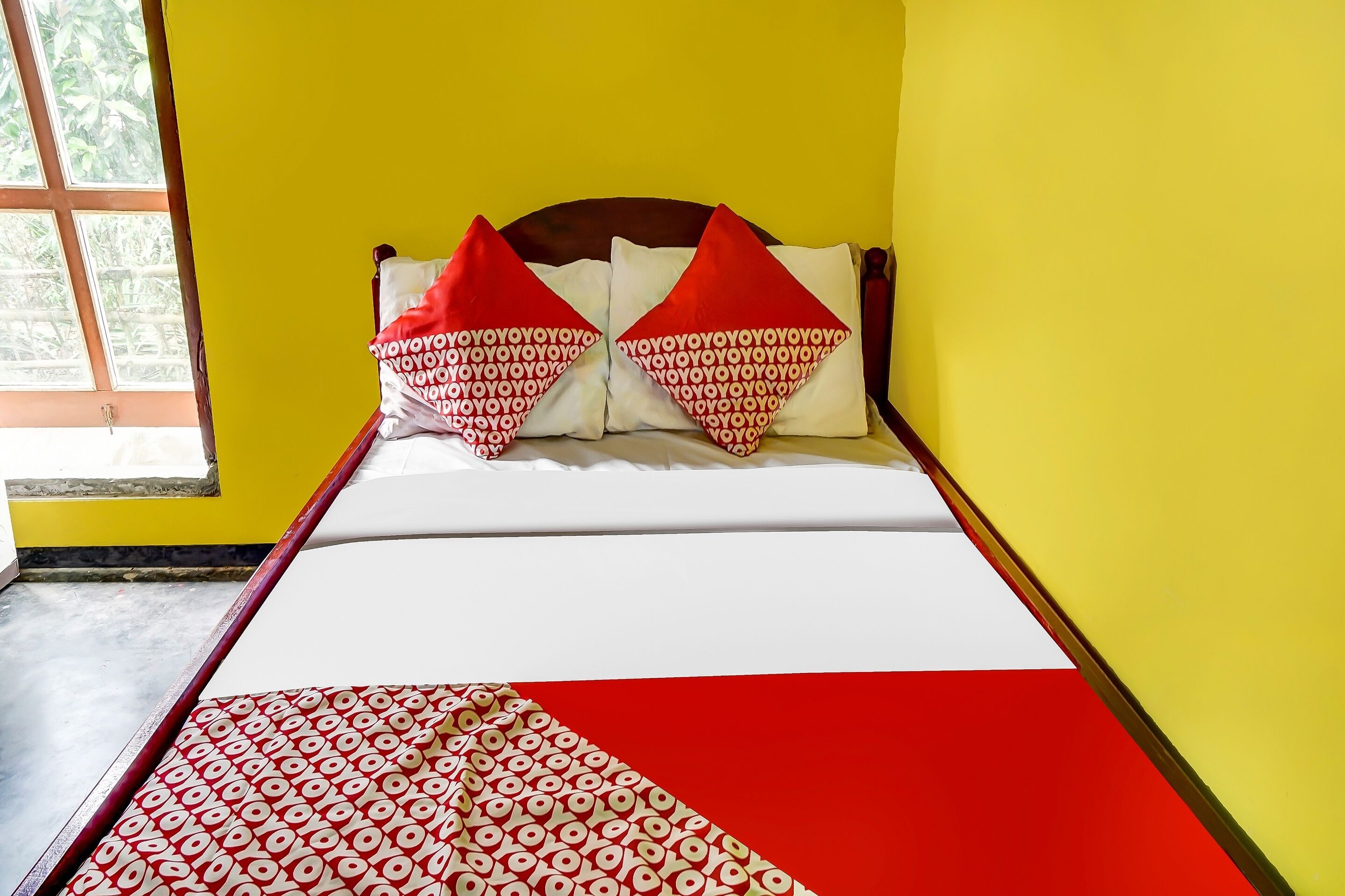 Un lit recouvert d’une couvre-lit à motifs rouges et blancs, deux oreillers aux motifs rouges et blancs, et une fenêtre donnant sur un paysage verdoyant.