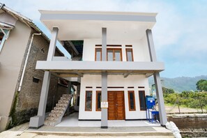 A two-story building with a white facade, a wooden door, and a staircase leading to the entrance. - OYO Homes 91008 Eco Tourism Desa Cibodas Babakan Gentong 1 Syariah (Lembang)