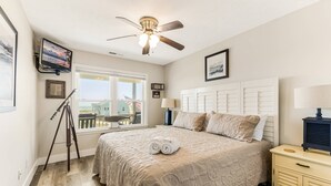 Une chambre à coucher avec un lit, un ventilateur de plafond, un téléviseur fixé au mur et une fenêtre donnant sur des immeubles.