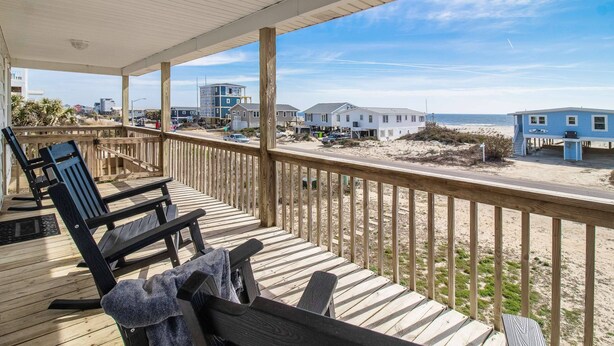 Une terrasse en bois, avec des chaises et un banc, donnant sur une plage bordée de maisons et sous un ciel dégagé.
