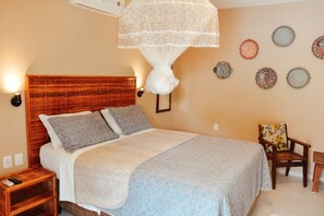 Un dormitorio con cabecera de madera, cama con ropa de cama gris y blanca, una mesita de noche con un control remoto, una silla con cojín floral y placas decorativas en la pared.