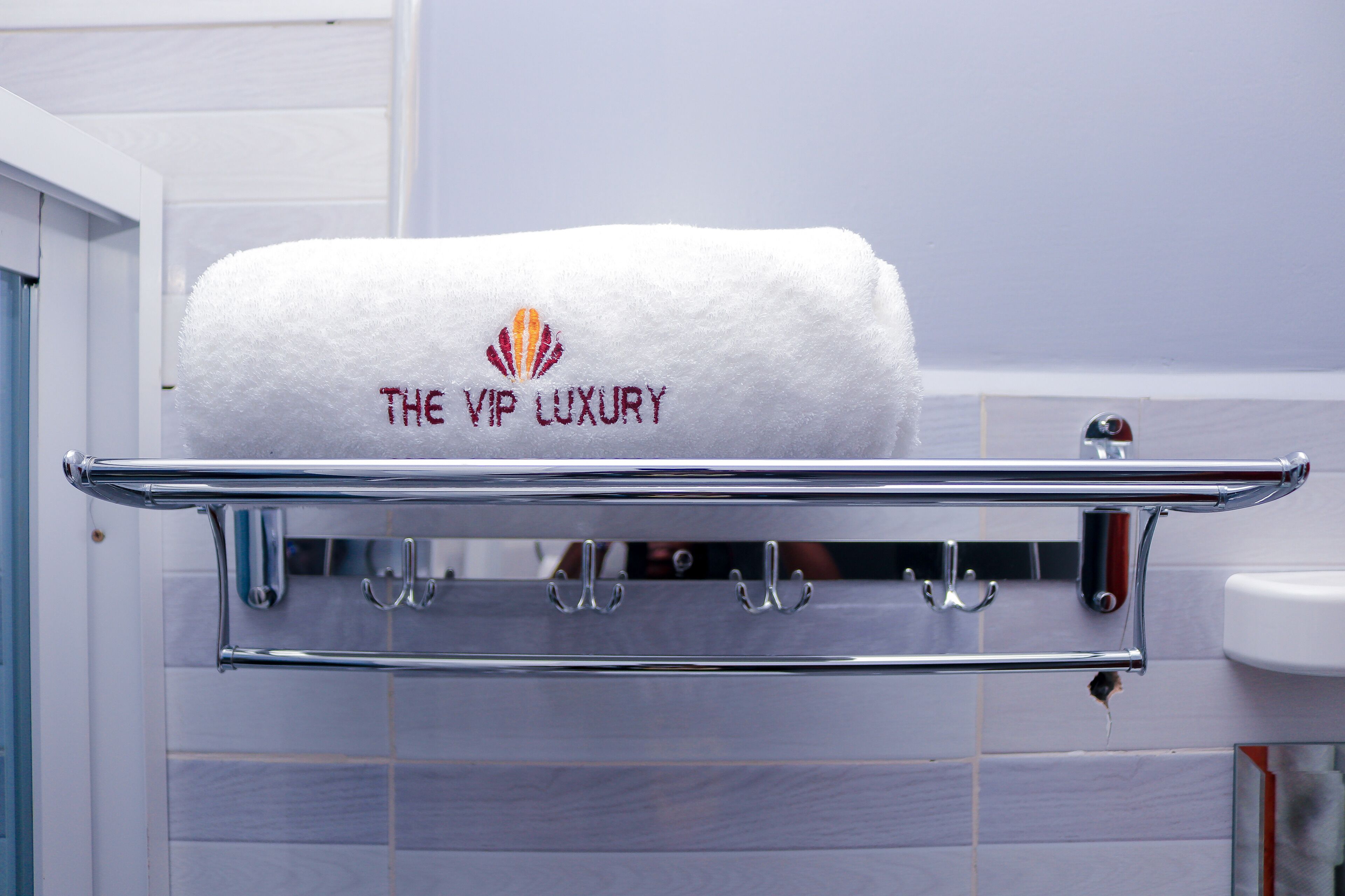 Une serviette blanche ornée d’une broderie « THE VIP LUXURY », suspendue à un porte-serviettes chromé dans une salle de bain aux murs carrelés.