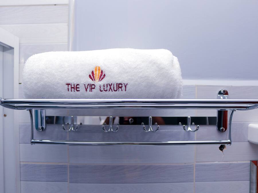 A white towel with "THE VIP LUXURY" embroidered on it, hanging on a chrome towel rack in a bathroom with tiled walls.