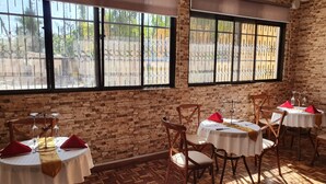 A restaurant with brick walls, large windows with metal bars, wooden flooring, and tables set with red napkins and glassware.
