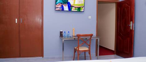 A hotel room with a bed, a desk with a chair, a television mounted on the wall, and a wardrobe.
