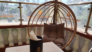 A wicker chair with a cushion, a black vase with a logo, and a view of a building through a glass railing.