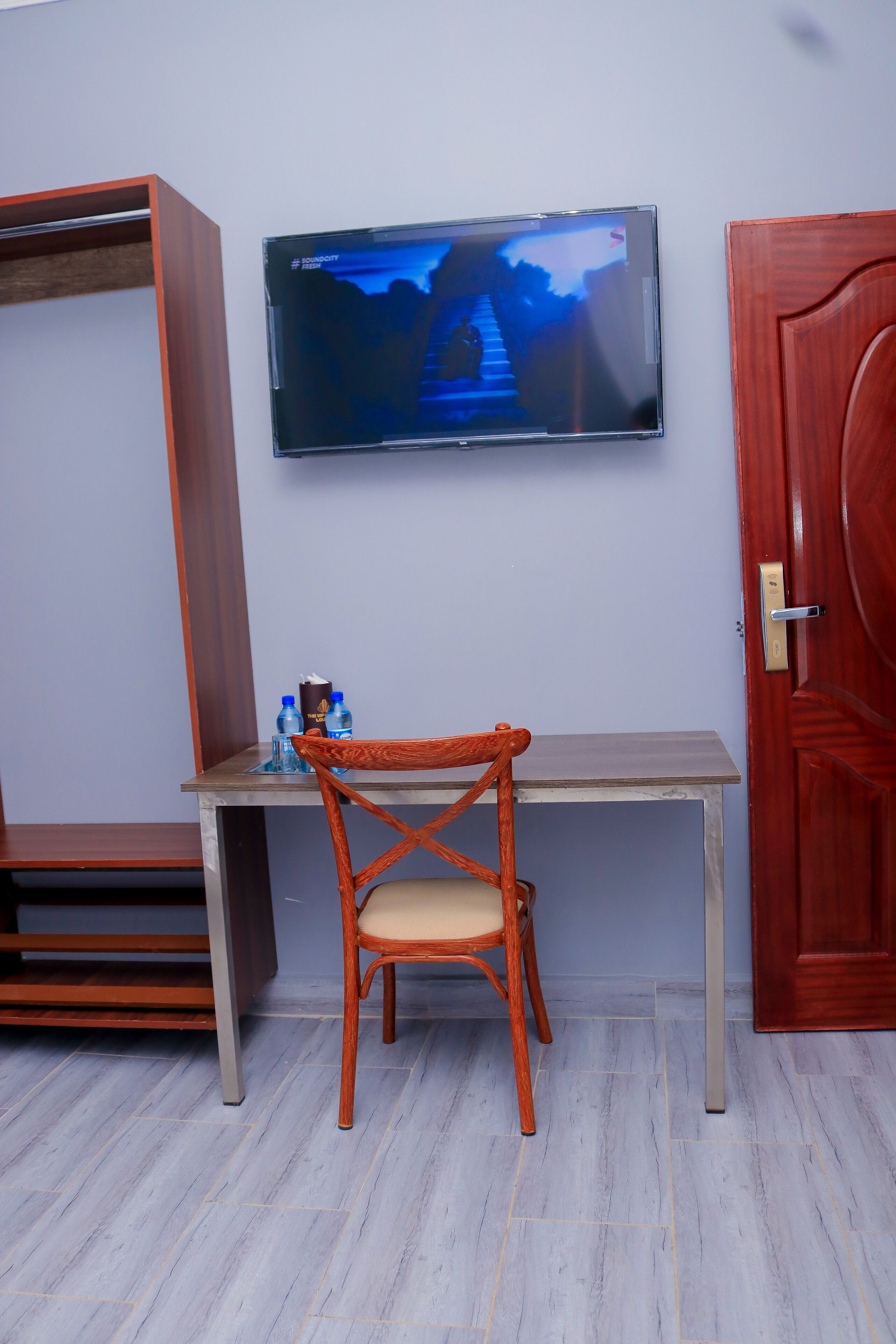 A room with a TV mounted on the wall, a wooden chair, a small table with water bottles, and a wardrobe.