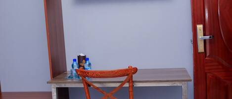 A room with a TV mounted on the wall, a wooden chair, a small table with water bottles, and a wardrobe.