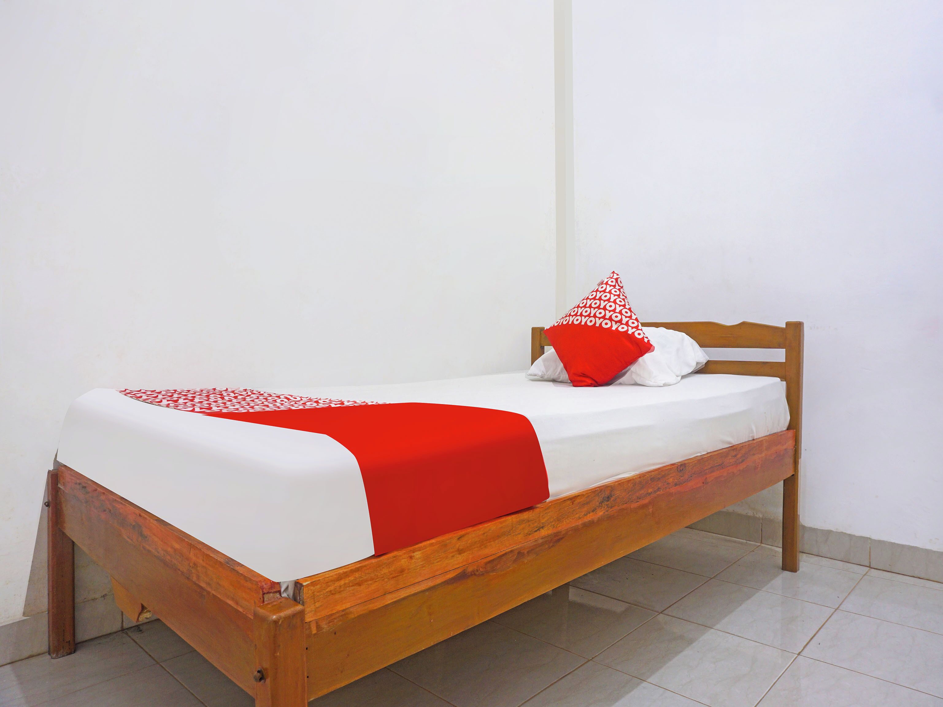 A single bed with a wooden frame, white bedding, and a red and white patterned pillow.