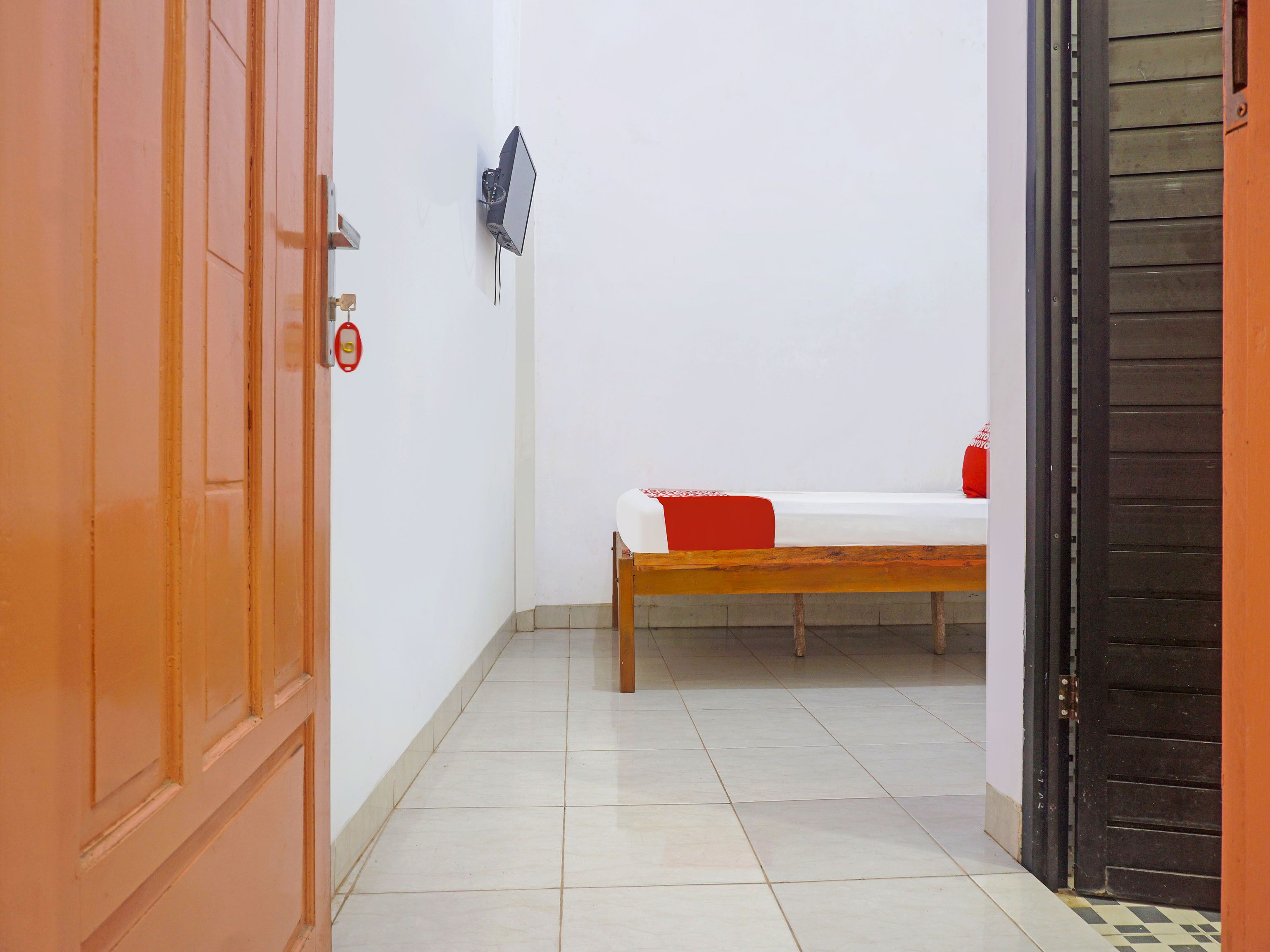 A narrow hallway with a bed in the background, a door on the left, and a wall-mounted device on the right.