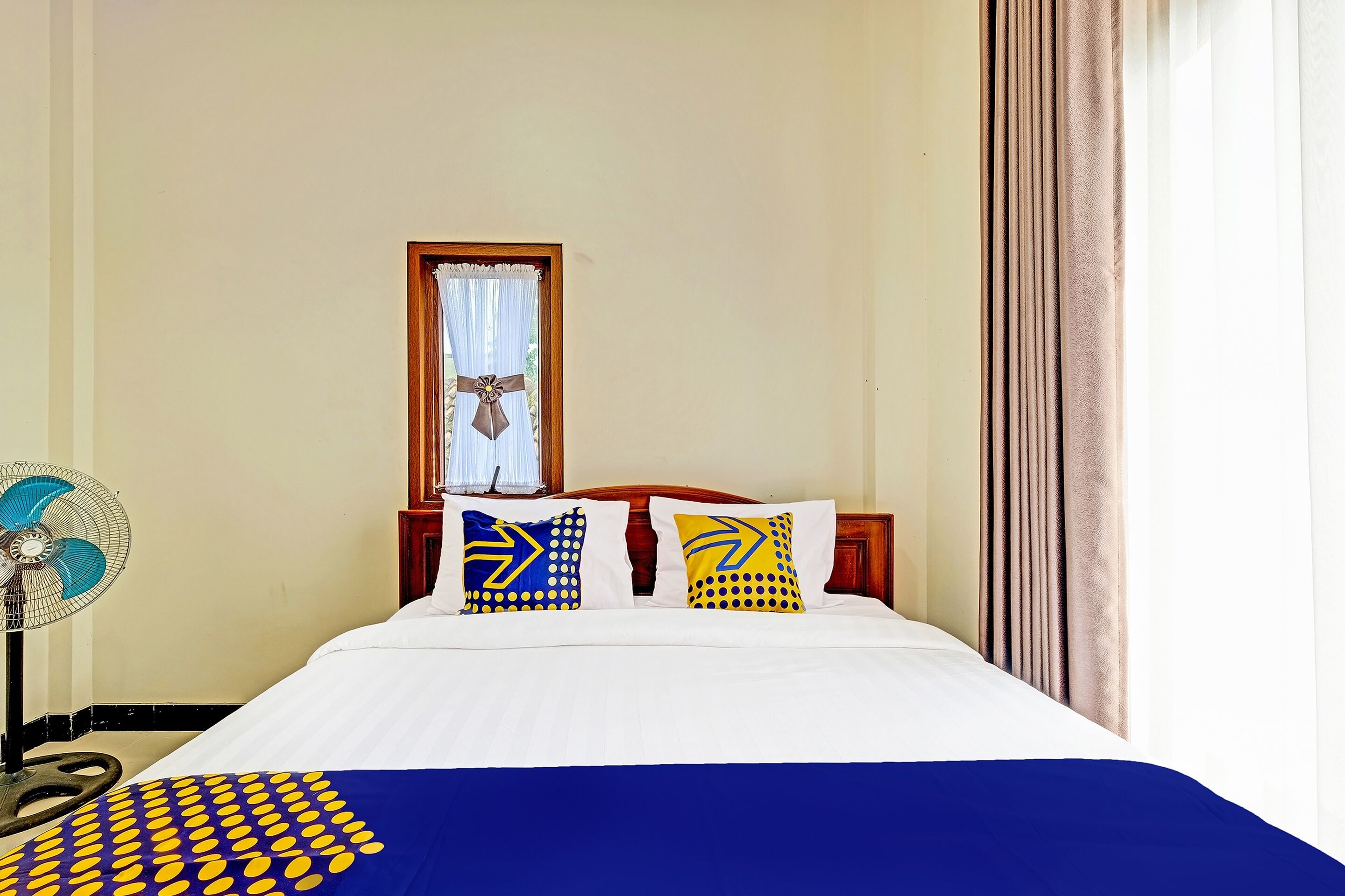 A hotel room with a bed, a fan, and a window with curtains.
