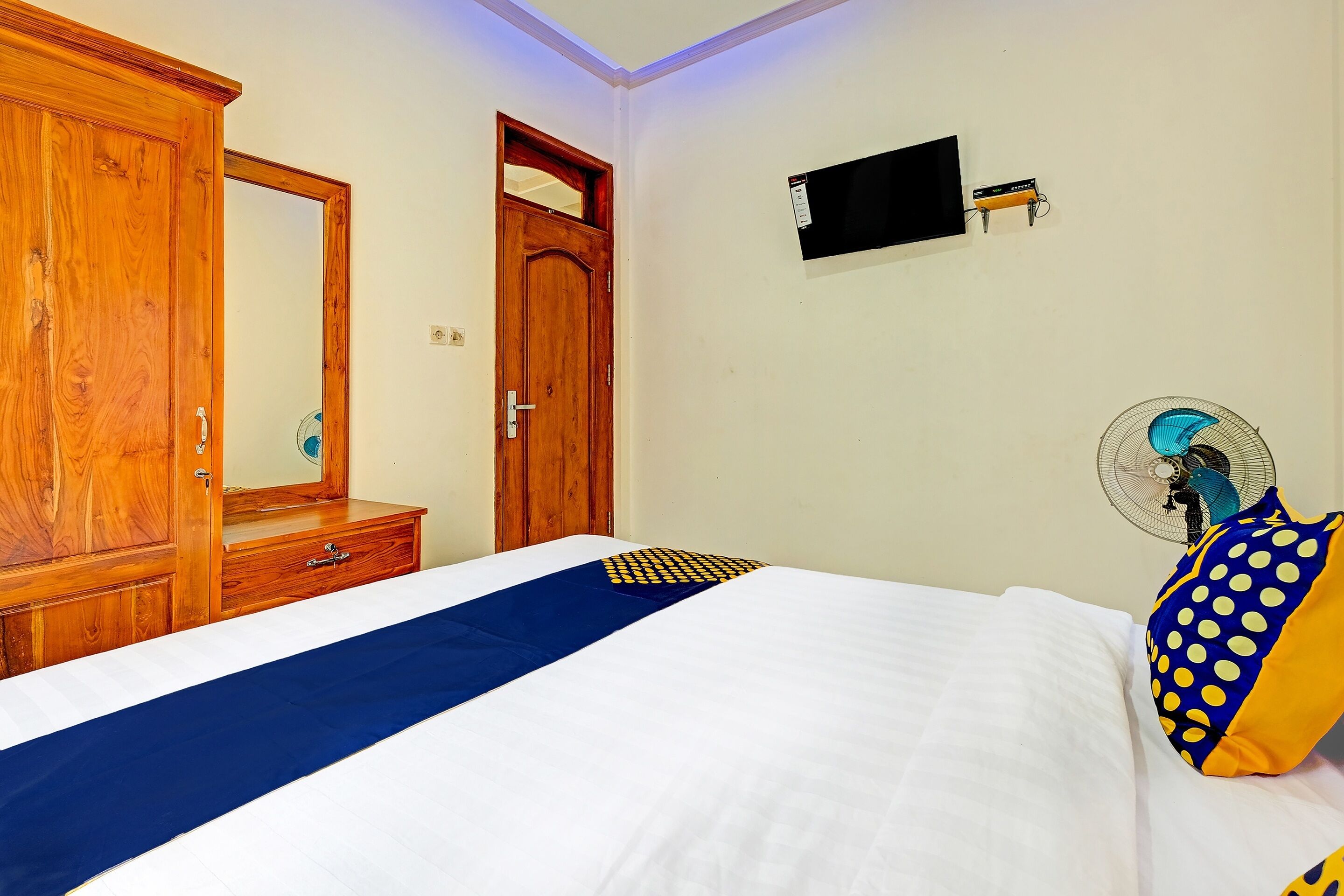 A bedroom with a wooden wardrobe, a bed with white bedding, a blue runner, a blue and yellow pillow, a wall-mounted TV, and a standing fan.
