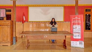 Eine Person sitzt an einem Holztisch mit einem Laptop in einem Raum mit Fliesenboden, roter Wand und einer großen Whiteboard-Tafel.