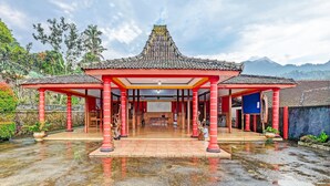 Ein traditionelles Pavillon mit einem Ziegeldach und roten Säulen, umgeben von einem spiegelnden, nassen Boden, mit Bergen im Hintergrund.