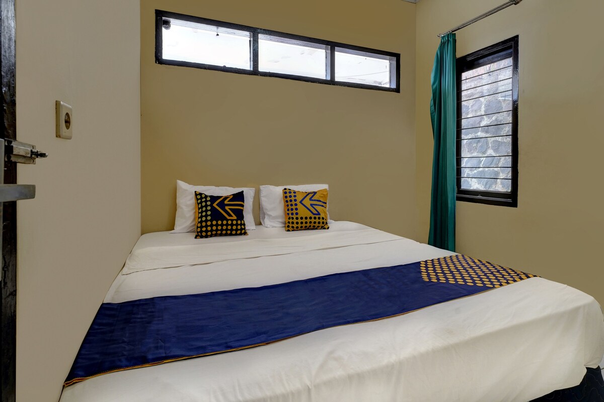 Ein Bett mit einer blauen und weißen Tagesdecke, zwei Kissen mit geometrischen Mustern, ein Fenster mit Jalousien und ein grüner Vorhang.