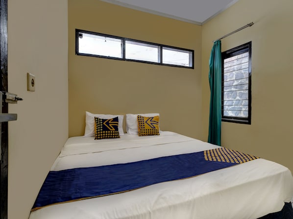 A neatly made bed with a blue and white bedspread, two pillows with geometric patterns, a window with a curtain, and a door with a handle.