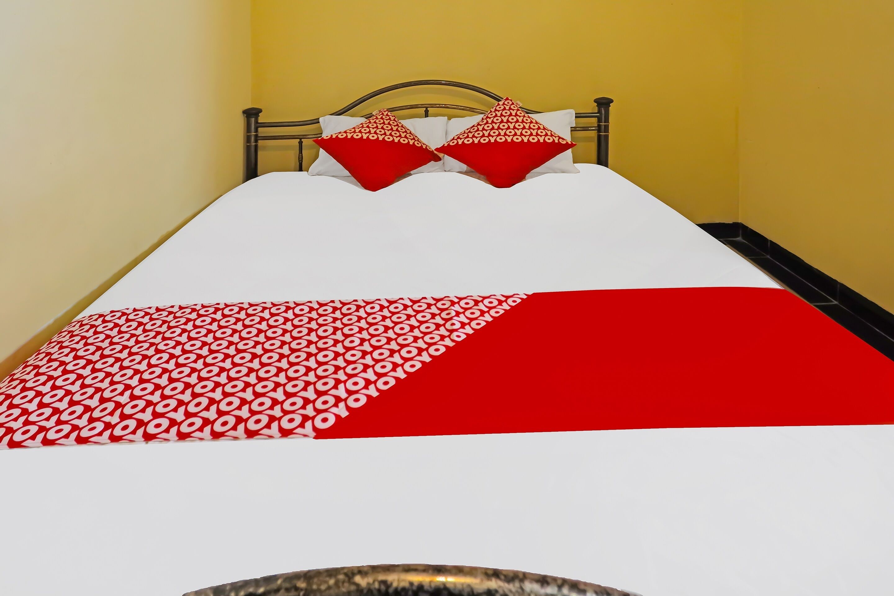 Een bed met een wit-rode spreideken, twee rode kussens met een cirkelpatroon, en een metalen hoofdbord.
